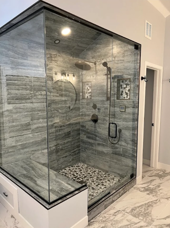 A modern glass-enclosed shower with gray stone-look tiles, a pebble floor, a built-in bench, and black hardware.