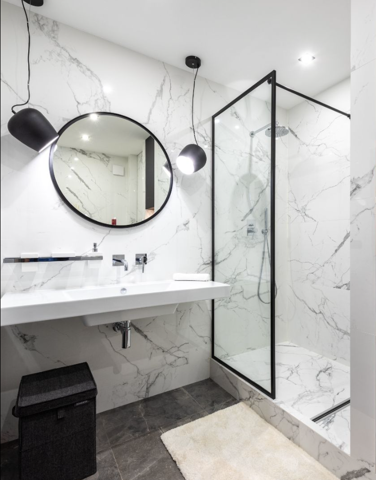 Modern bathroom with a marble wall, round mirror, black pendant lights, a floating sink, and a walk-in glass shower.