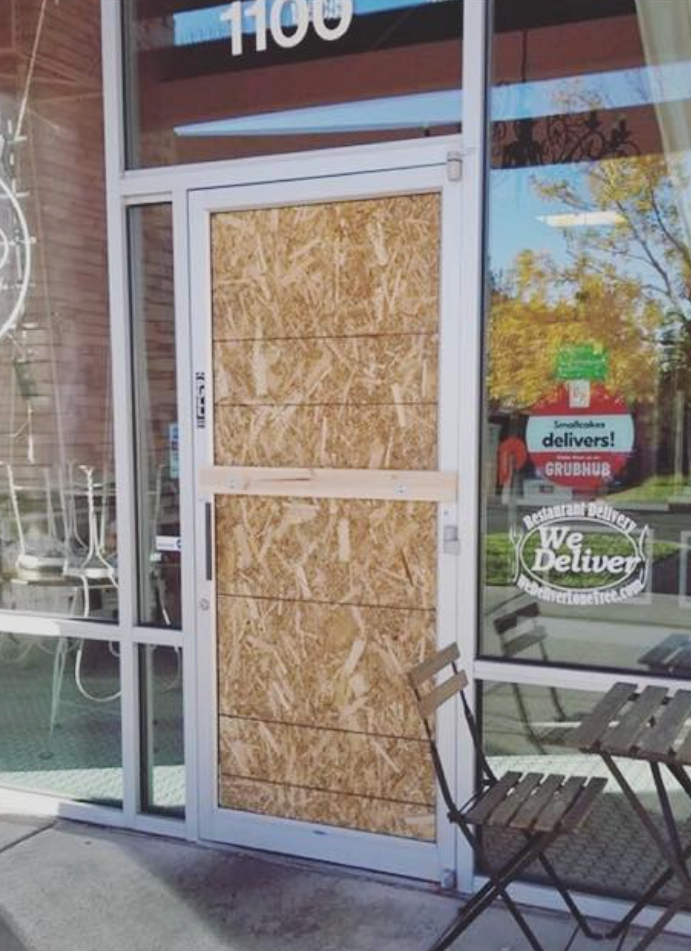 A door boarded up with wood panels at a business storefront; outdoor seating nearby.