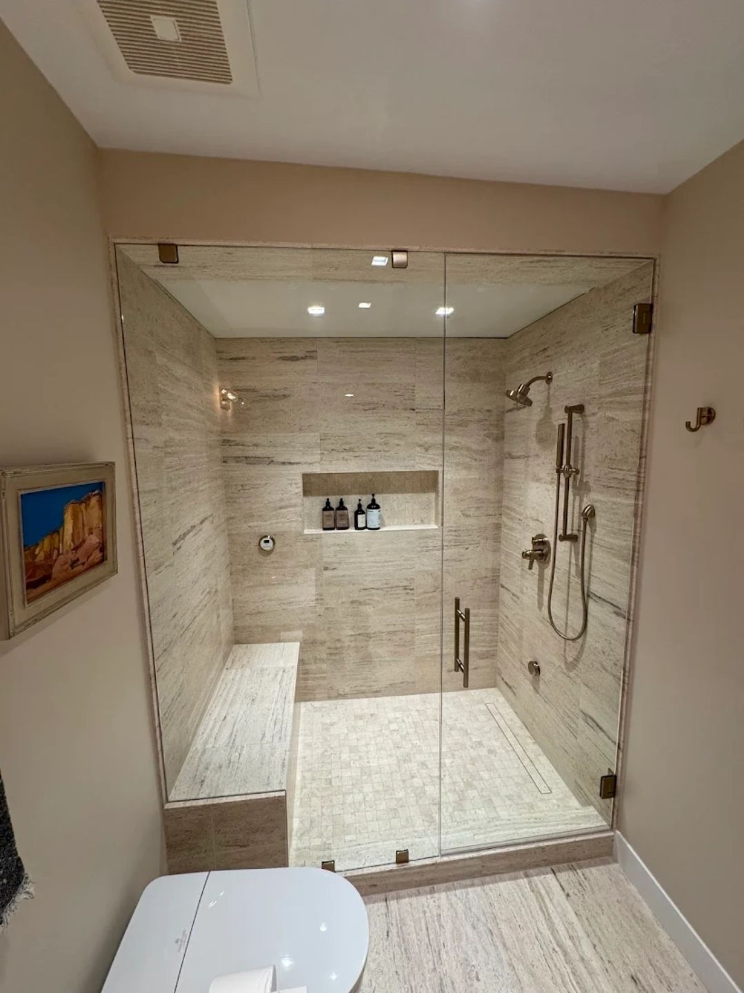 A modern walk-in shower with beige marble tiles, a built-in bench, a glass door, and a wall-mounted shower head.