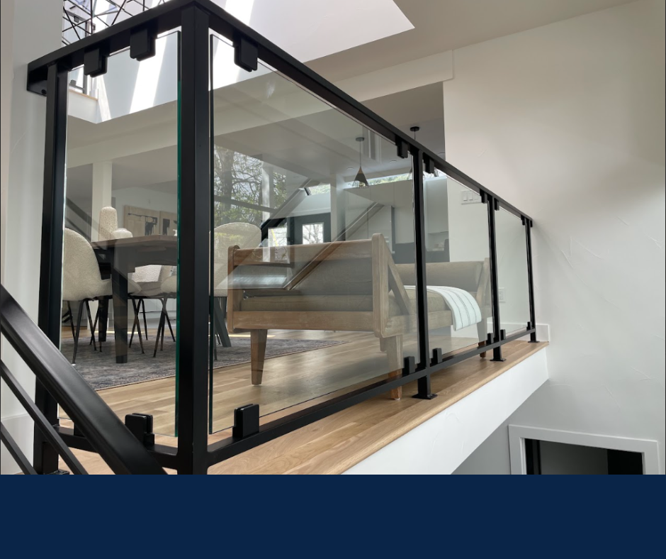 A black metal railing with glass panels borders a wooden floor overlooking a bright living room.