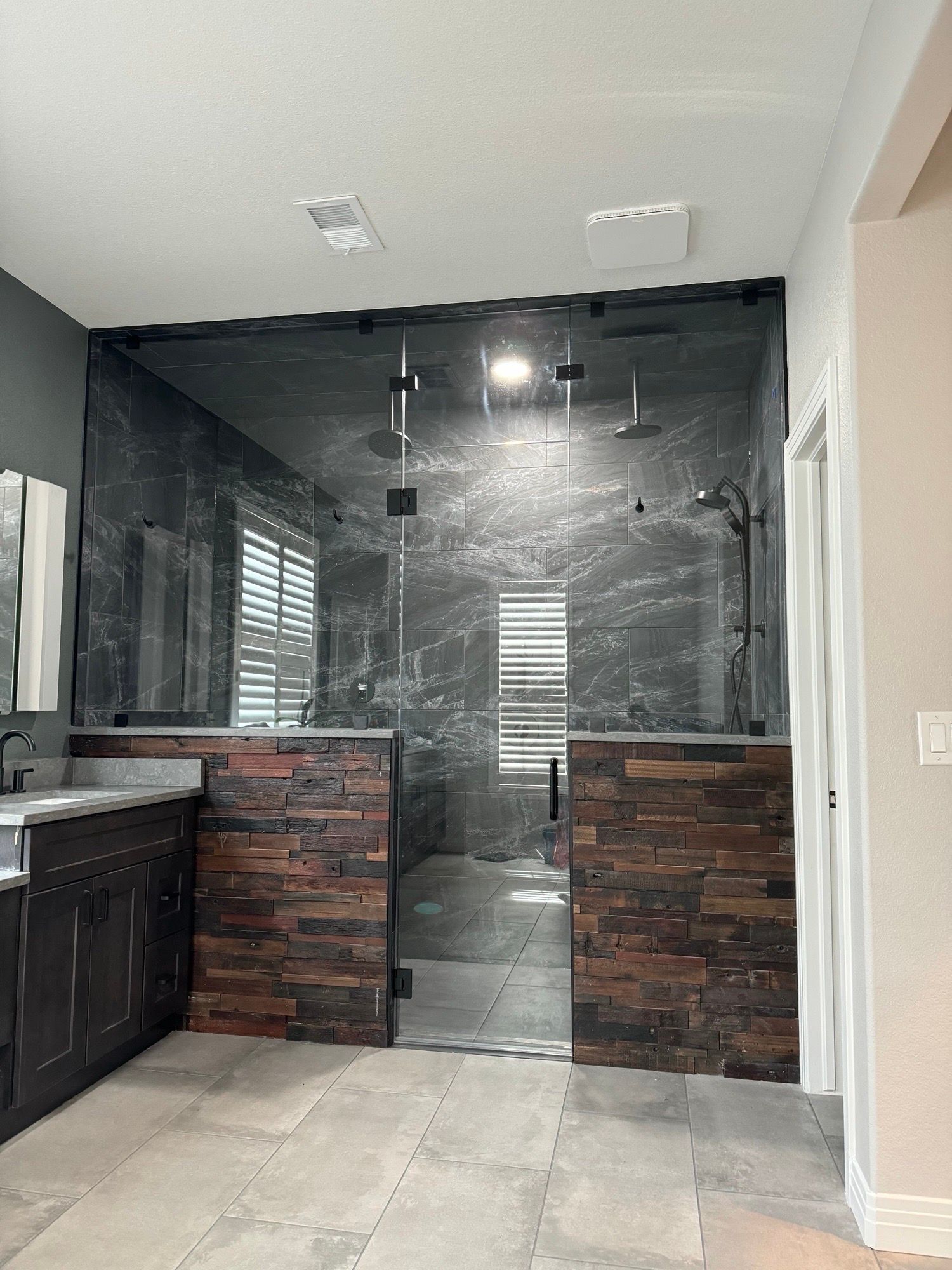 Modern bathroom with a glass-enclosed shower featuring dark stone tile walls, light tile floors, and a dark wood vanity.