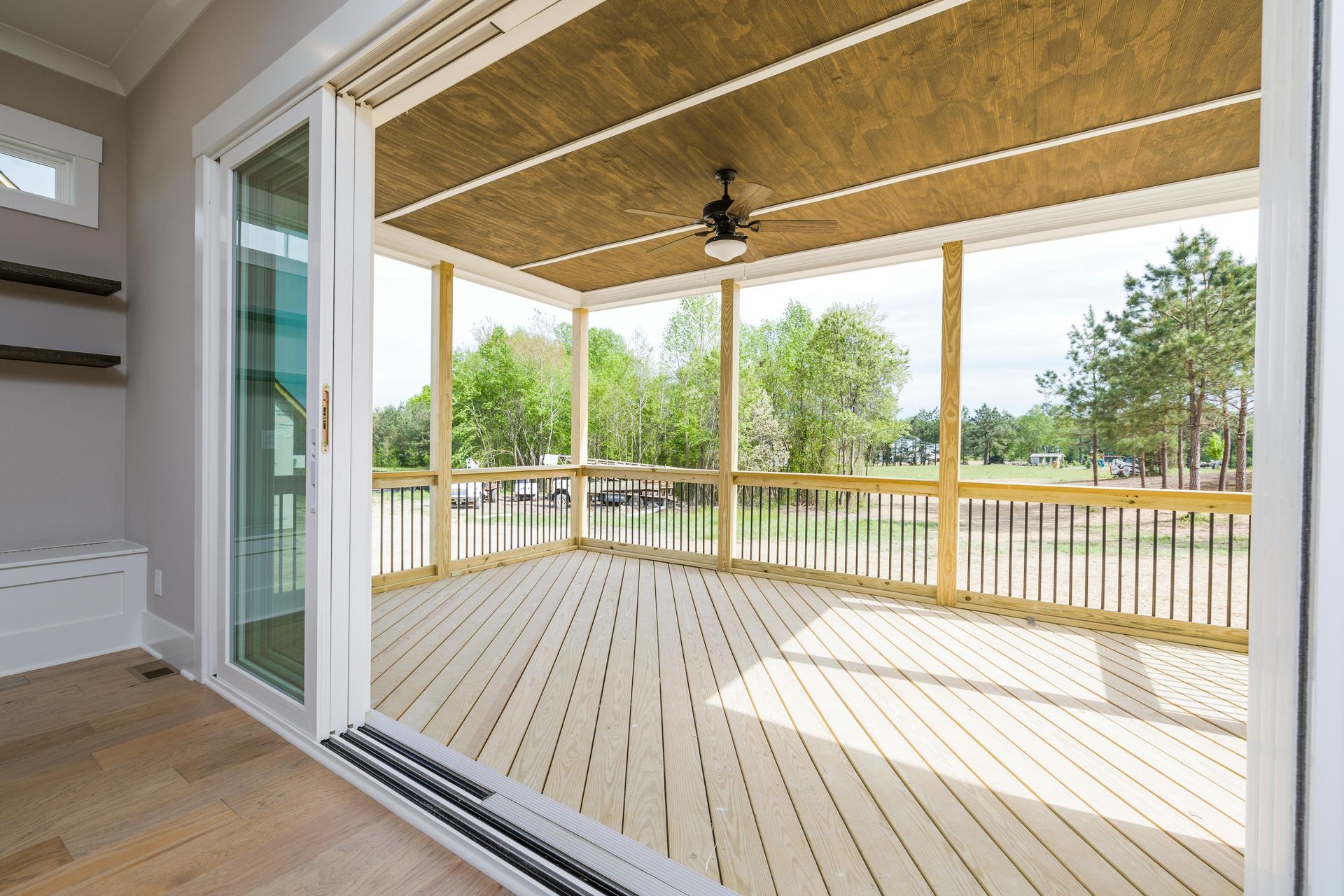 View of an open porch with wooden deck and railing, overlooking a yard with trees and cars.