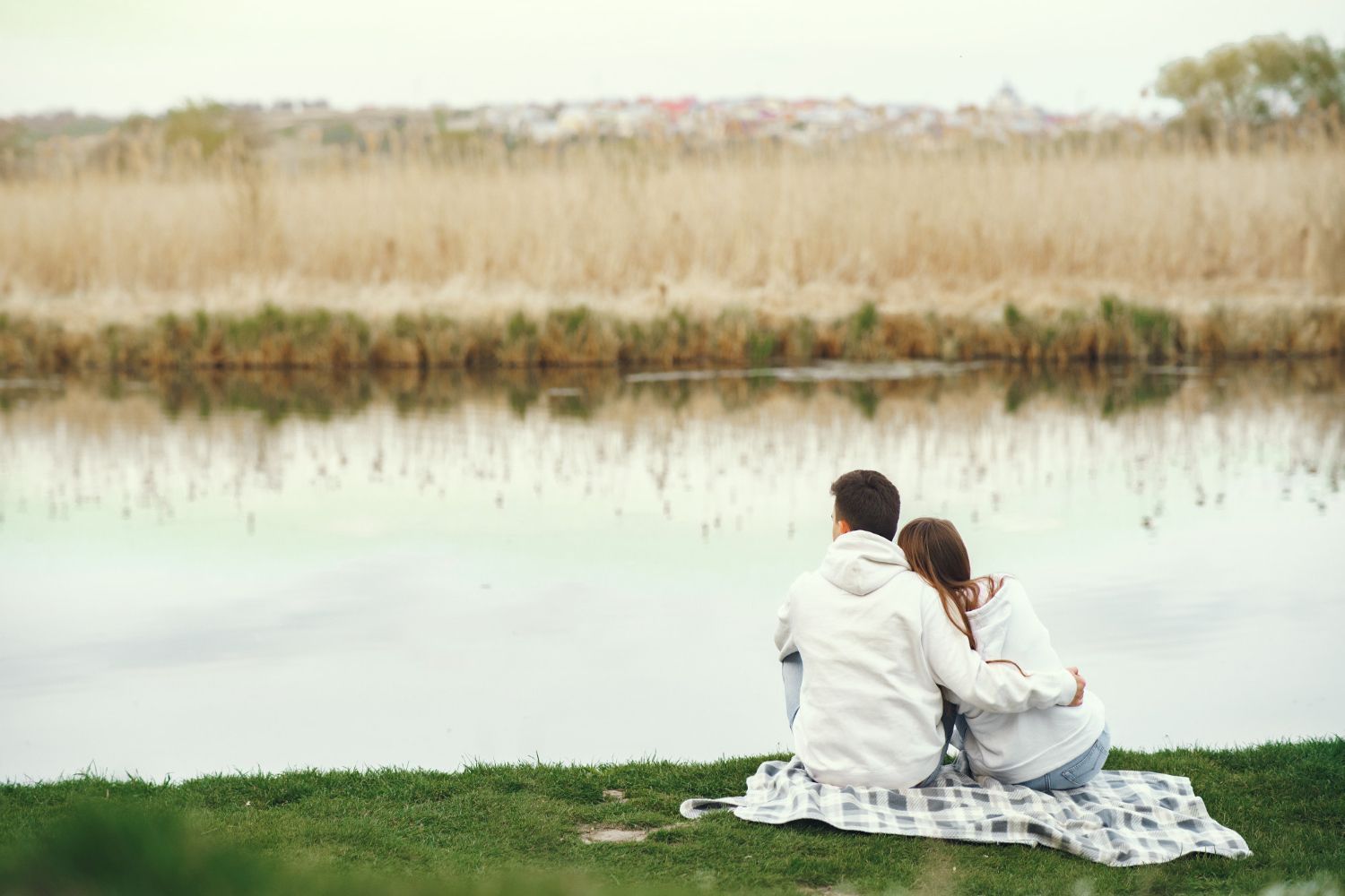 Couple seated on a blanket by a lake, looking at the water. Man has arm around woman. Reeds in background.