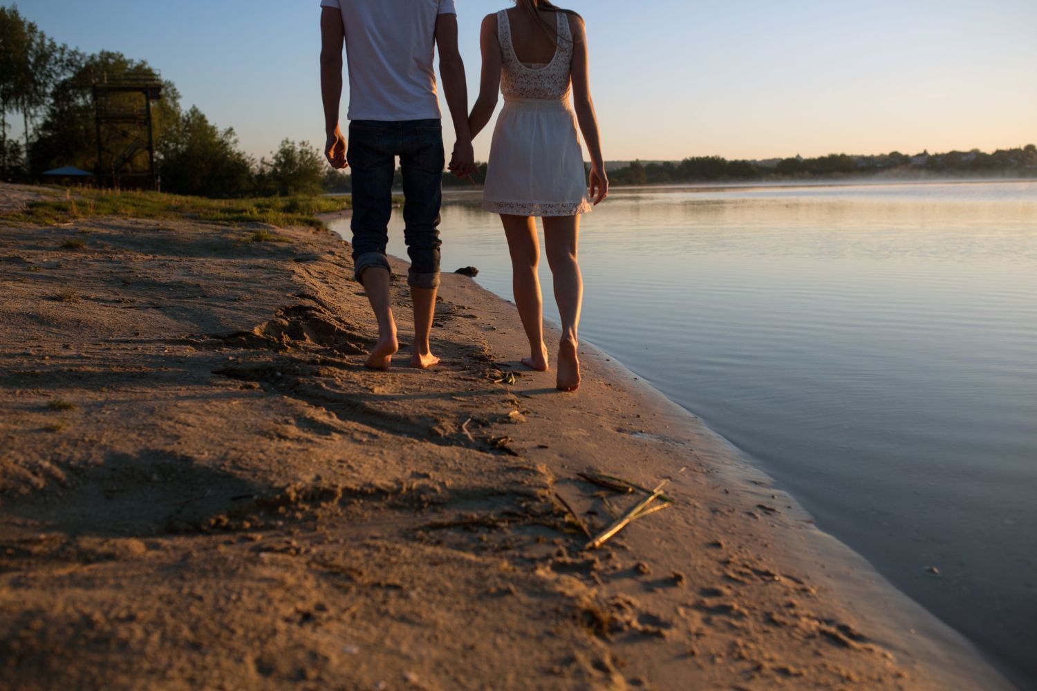 Couple, holding hands, walking barefoot along a sandy beach by a calm lake at sunset.