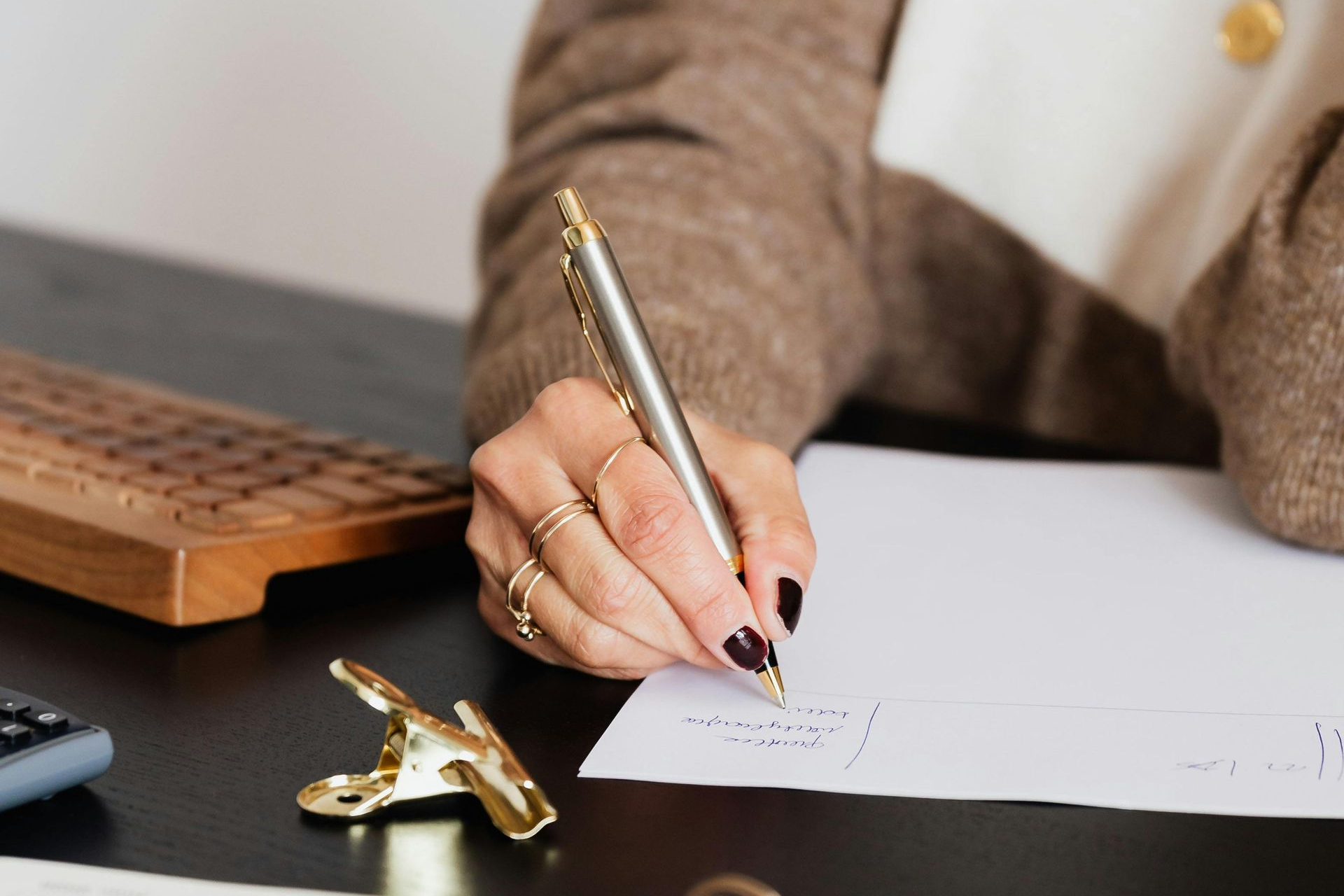 Woman writing on paper with a pen, rings on her fingers, black nail polish, at a desk.