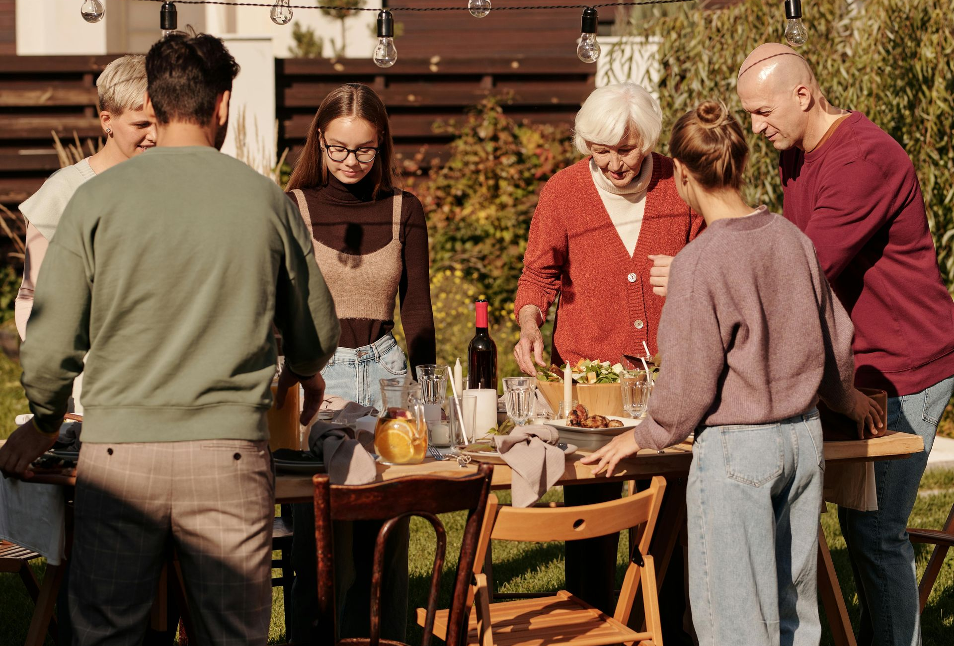 Family gathers around a table outdoors, preparing and serving food. Sunny setting.
