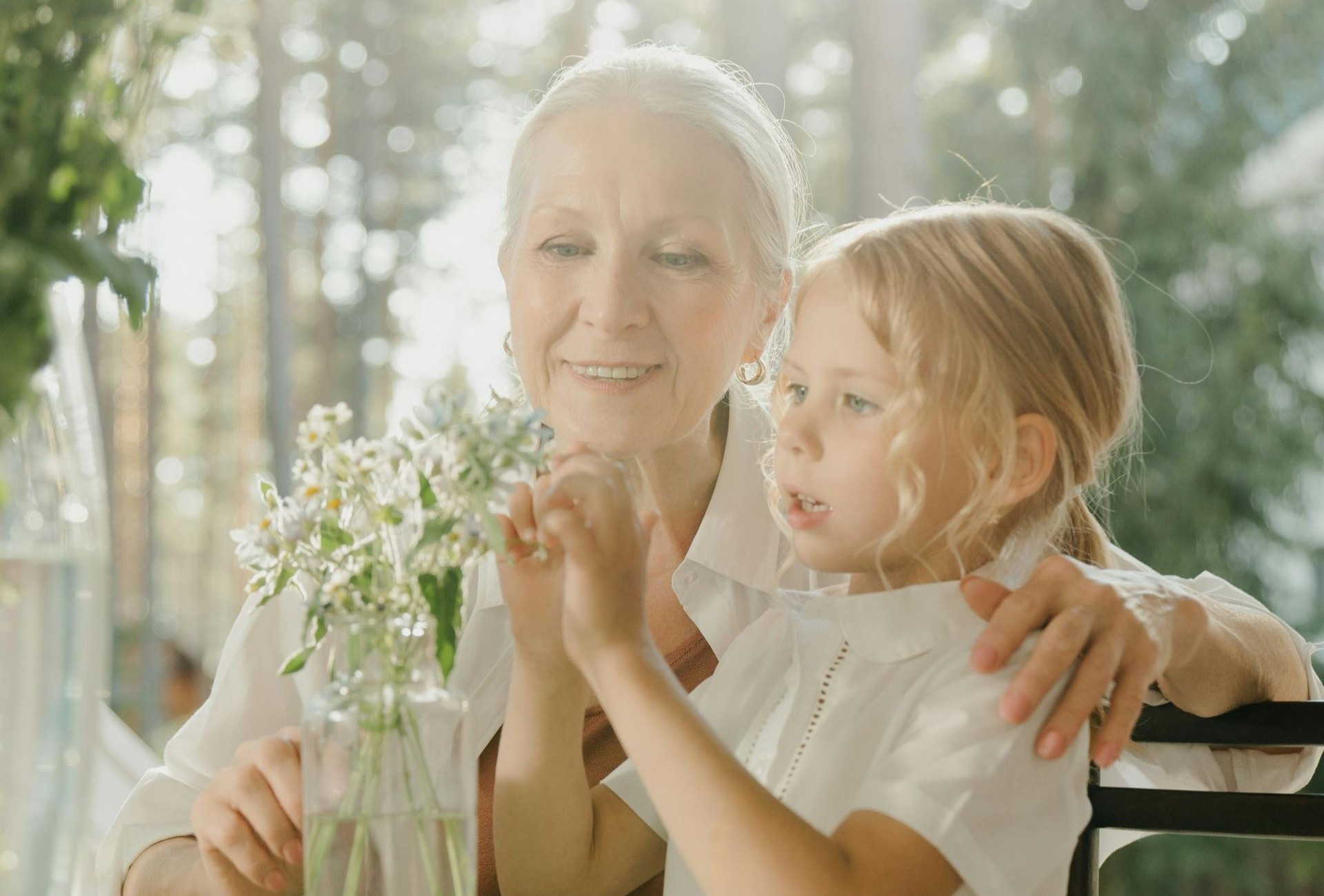 Grandmother with a young girl, seated at a table with flowers, smiling, sunny outdoor setting.