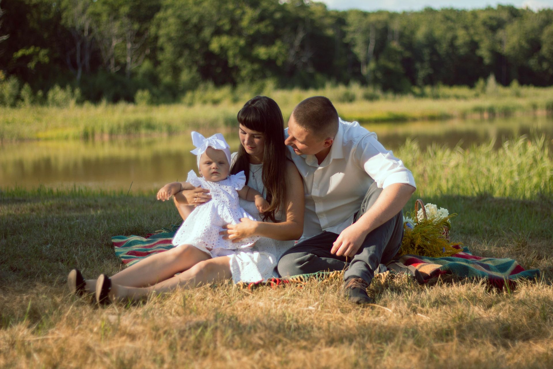 Family of three on a picnic blanket by a lake, the child in a white dress and the parents smiling.