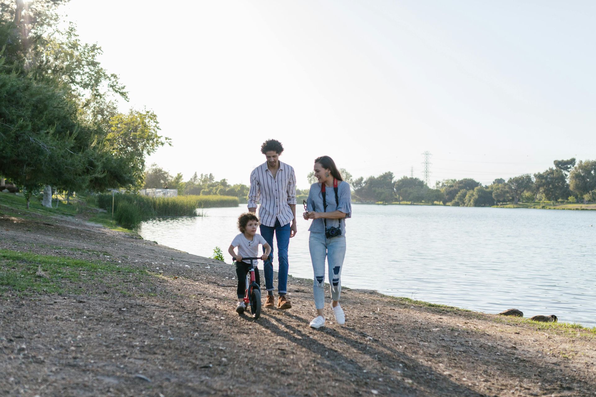 Family walking along a lake, child on scooter, mother with prosthetic leg, sunny day.