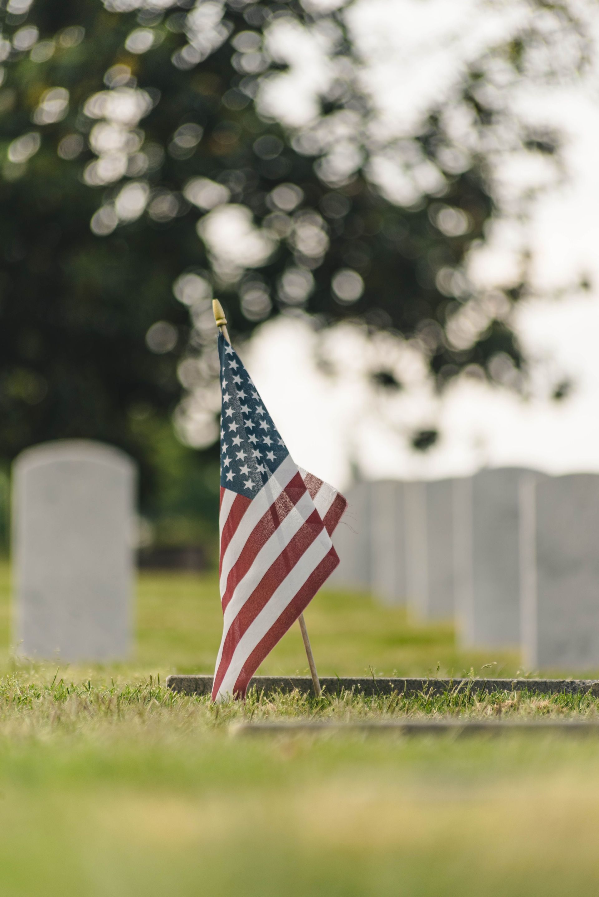 Small American flag planted in grass at a cemetery with blurred headstones in the background