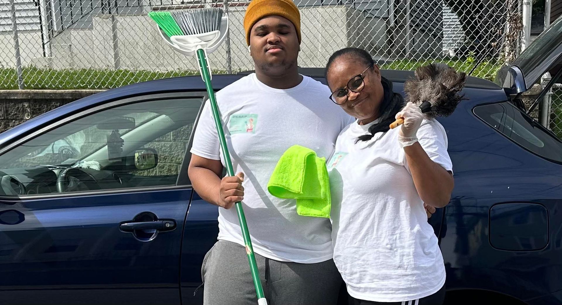 Two people stand outside, smiling and holding cleaning supplies.