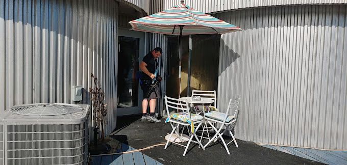 A man is standing on a patio with a table and chairs under an umbrella.