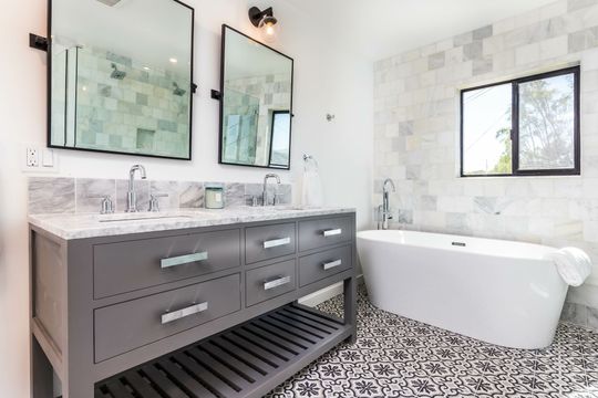 A bathroom with a gray double vanity, marble countertops, two rectangular mirrors, and a white freestanding bathtub.