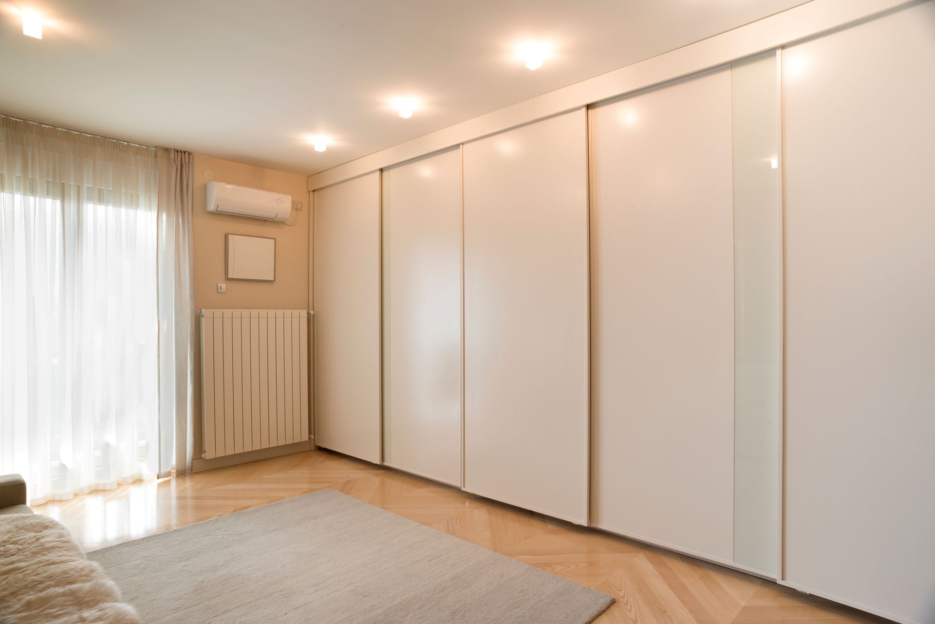 A bright room features a large white wardrobe, a neutral area rug, parquet flooring, and curtains covering a window.