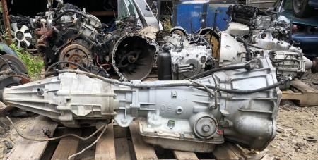 Automatic transmission on a wooden pallet, surrounded by other vehicle parts in a junkyard.