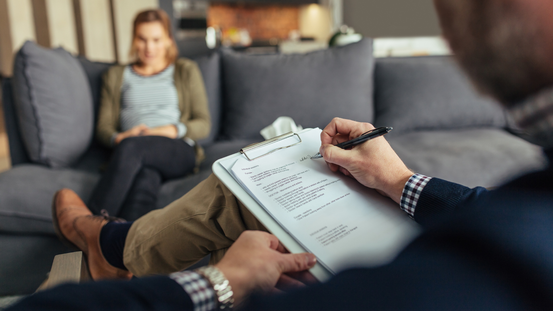 A man is writing on a clipboard while a woman sits on a couch.