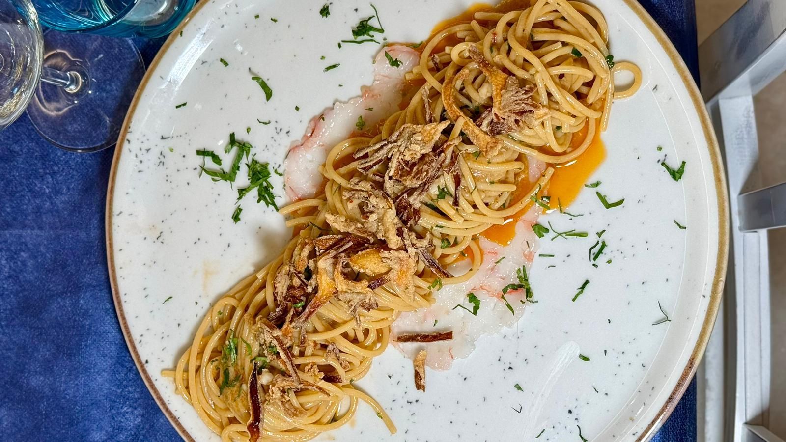 Piatto di pasta servito su un piatto bianco con condimenti fritti e guarnizione su una tovaglia blu.