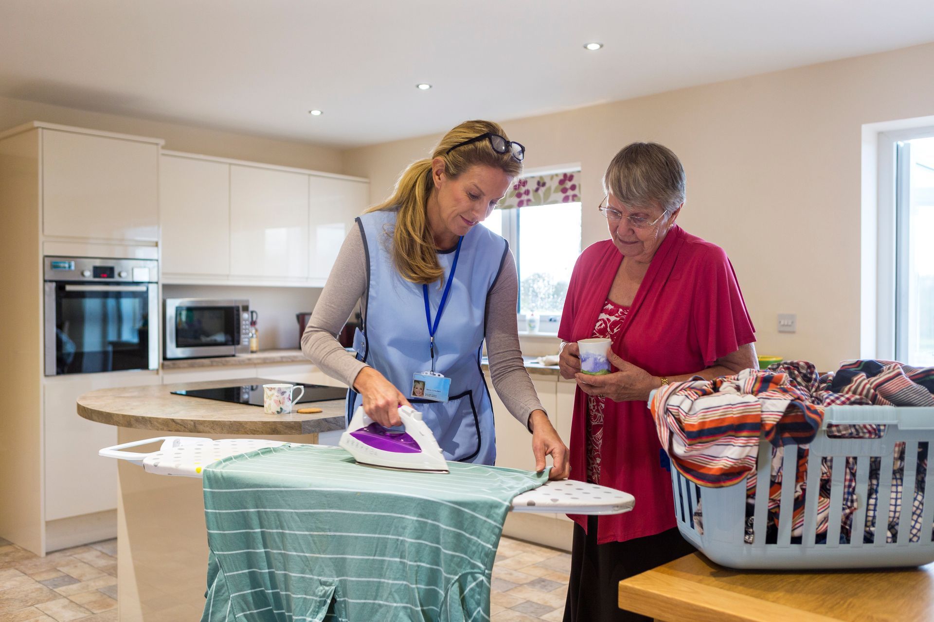 A woman is ironing a shirt in a kitchen while another woman looks on.