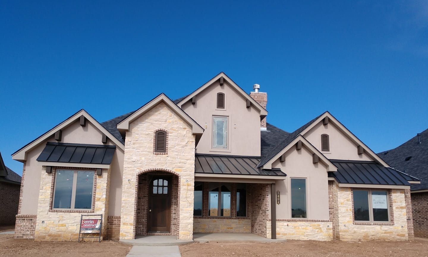 Two-story house with tan stucco and stone facade, black metal roof, and blue sky.