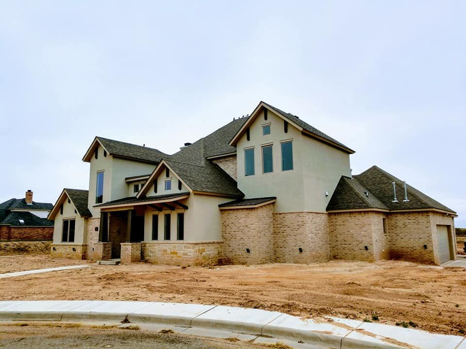 Large beige house with brick and tan stone accents, built on a dusty lot under a cloudy sky.