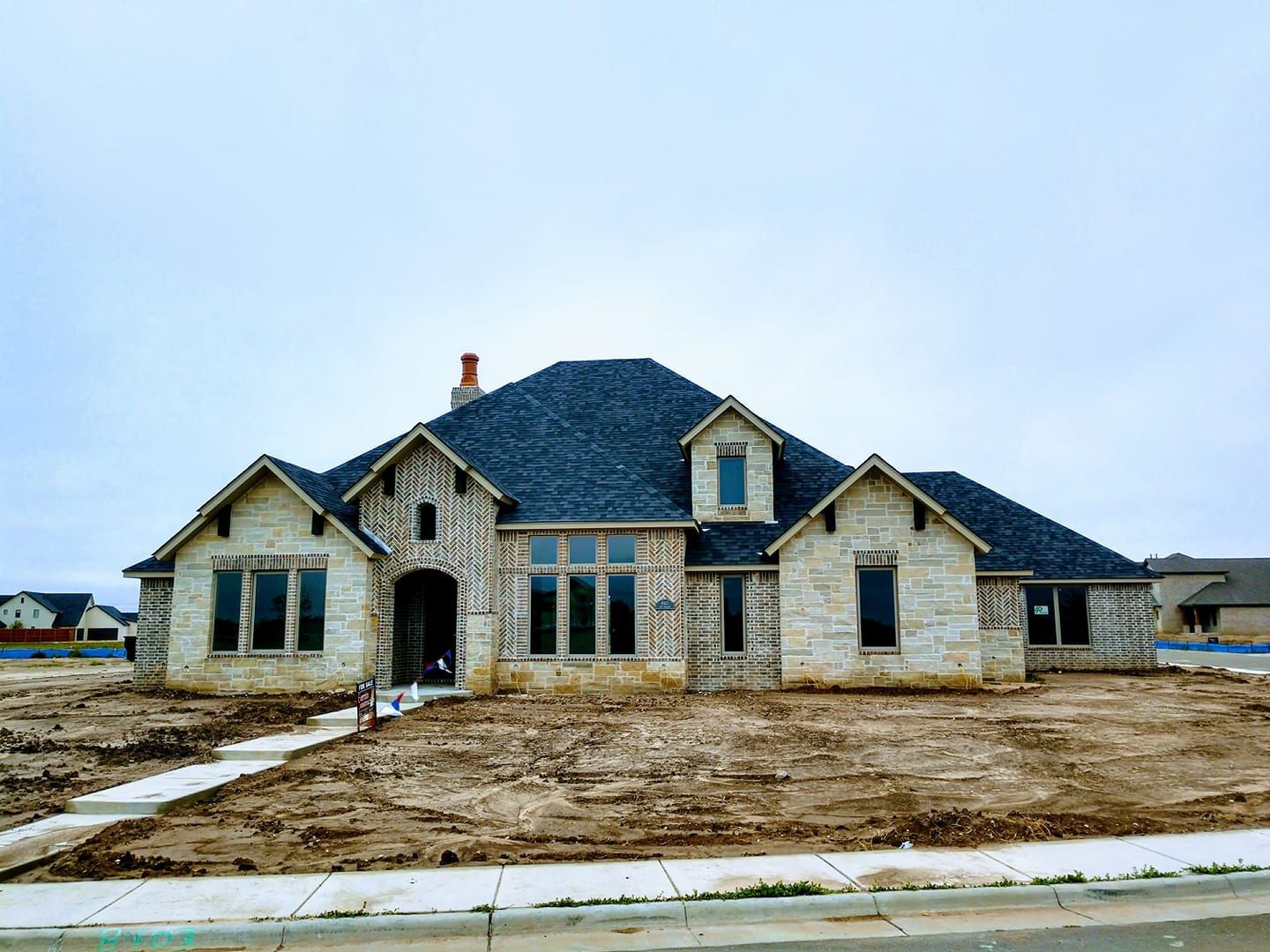 New house under construction with stone facade, brick details, and dark roof on a cloudy day.