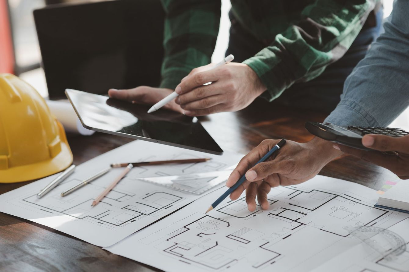 People reviewing blueprints, using tablet and pens, with a yellow hard hat on a table.