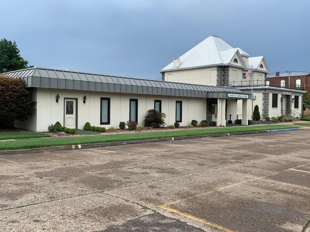 Cream-colored building with dark roof and trim. Small bushes line the front. Cloudy sky.