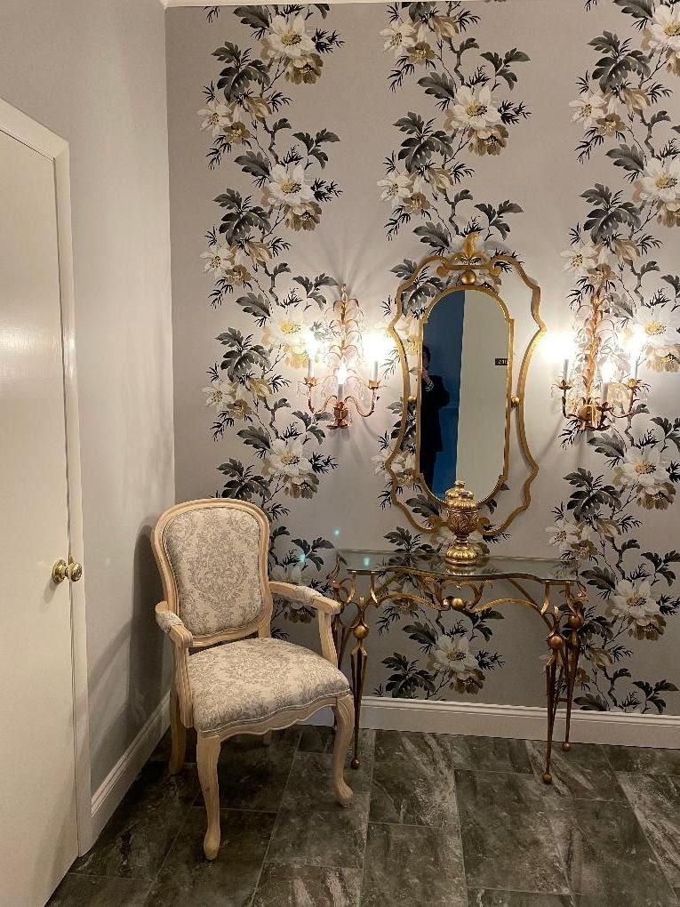 A hallway with floral wallpaper, ornate mirror, gold table, sconces, and a chair.