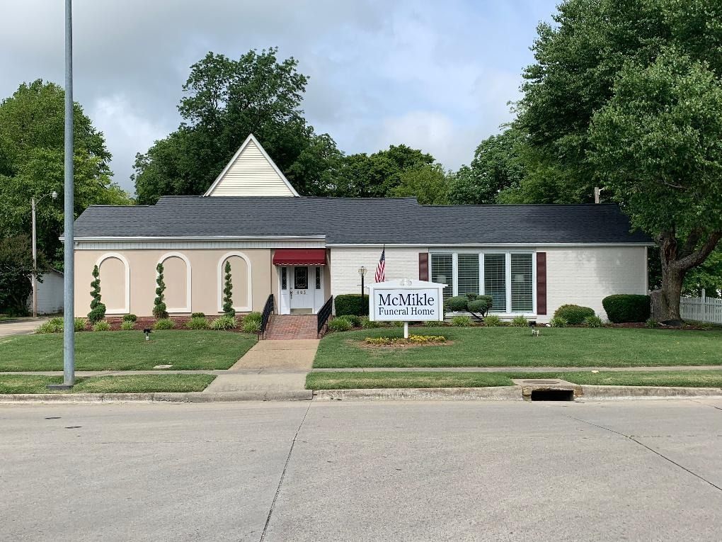 McVay Home of Funerals building, white brick, black roof, red awning, green lawn, sign, and trees.