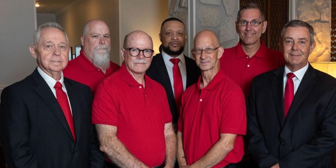 Group of men in red shirts and suits posing in front of a neutral background.