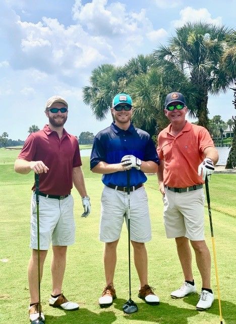Bill Lovell and his two sons posing on a golf course green with clubs, palm trees, and a bright cloudy sky behind them.