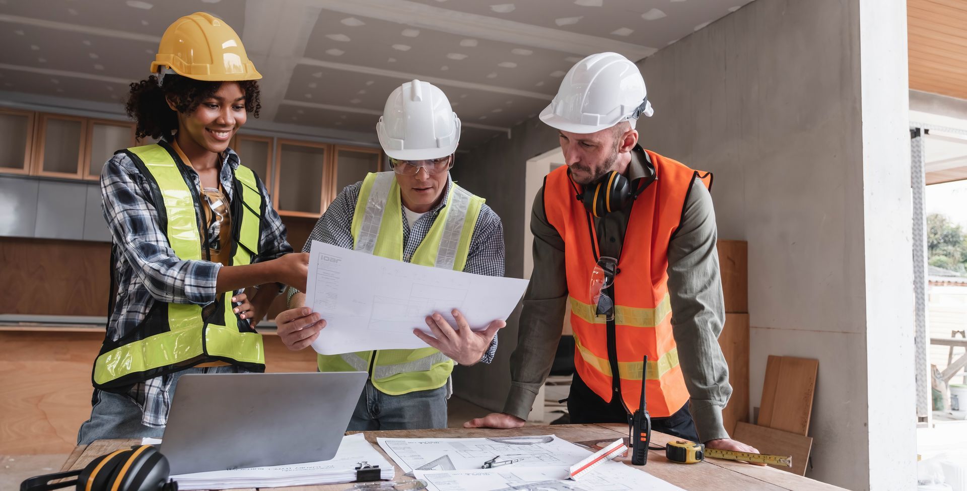 Three construction workers in safety gear reviewing blueprints at a building site