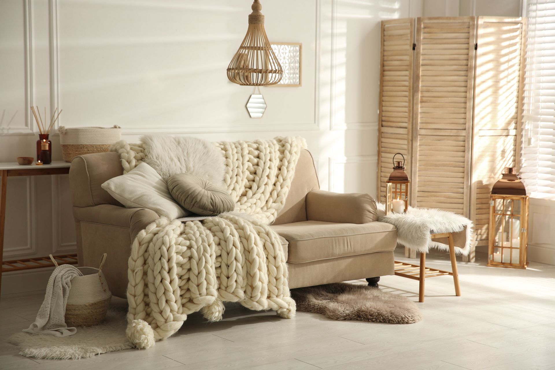 Cozy beige living room with a sofa, chunky knit throw, woven lamp, and rattan screen in soft sunlight