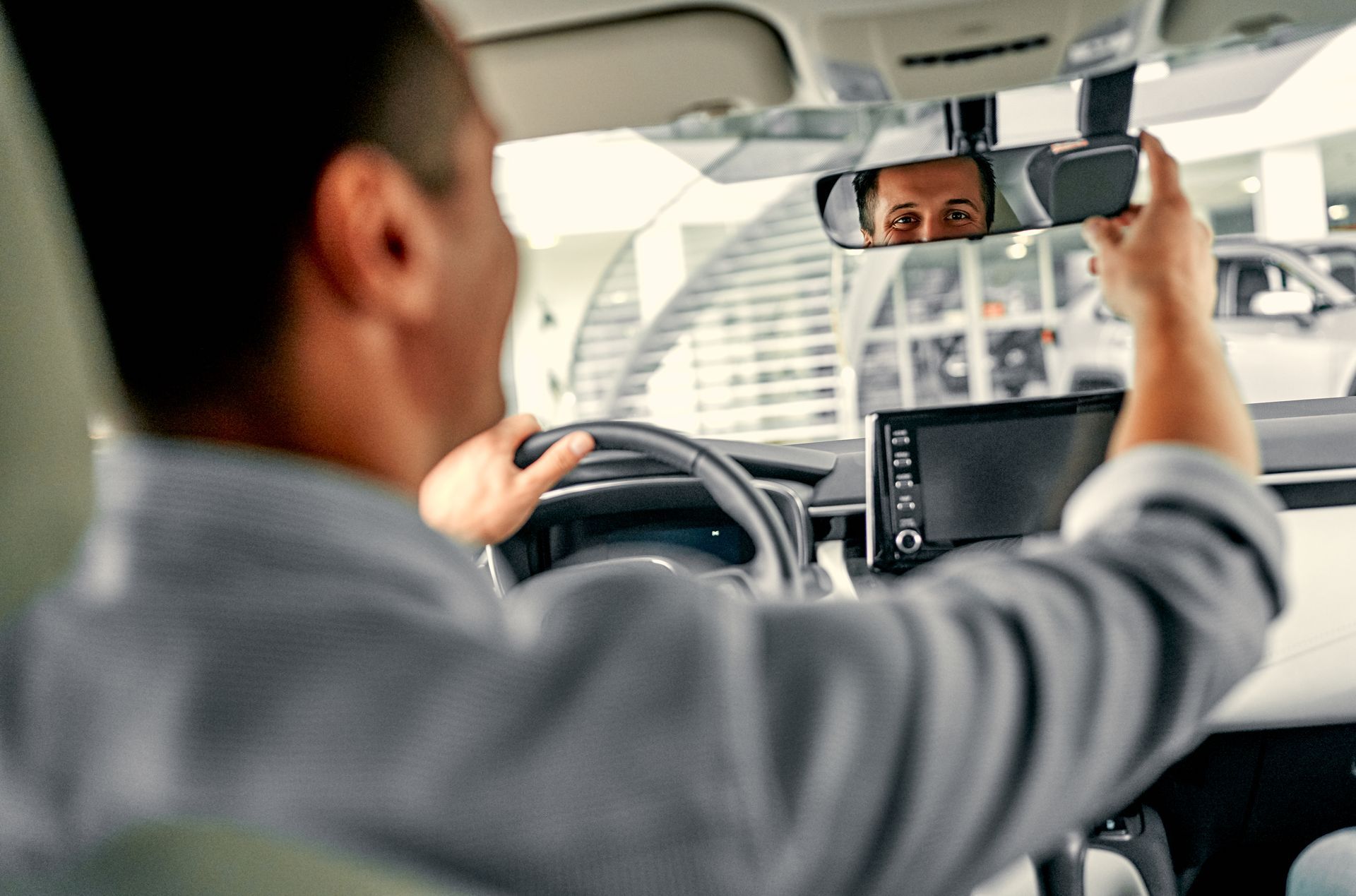 Driver adjusting rearview mirror in a car, with dashboard screen and city buildings visible ahead