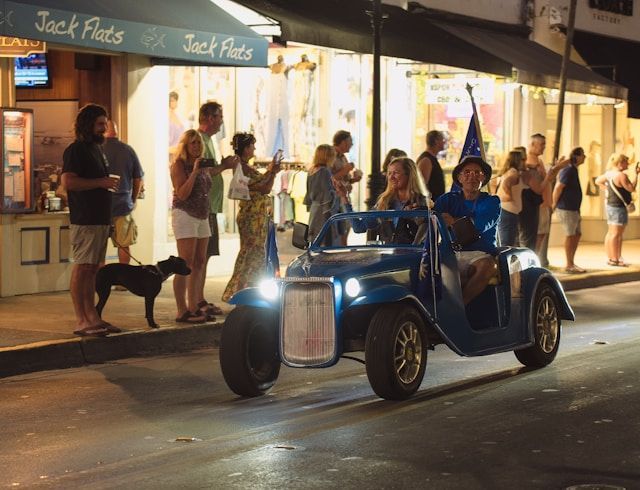 Blue vintage car driving past a bright storefront at night, with pedestrians watching on the sidewalk.