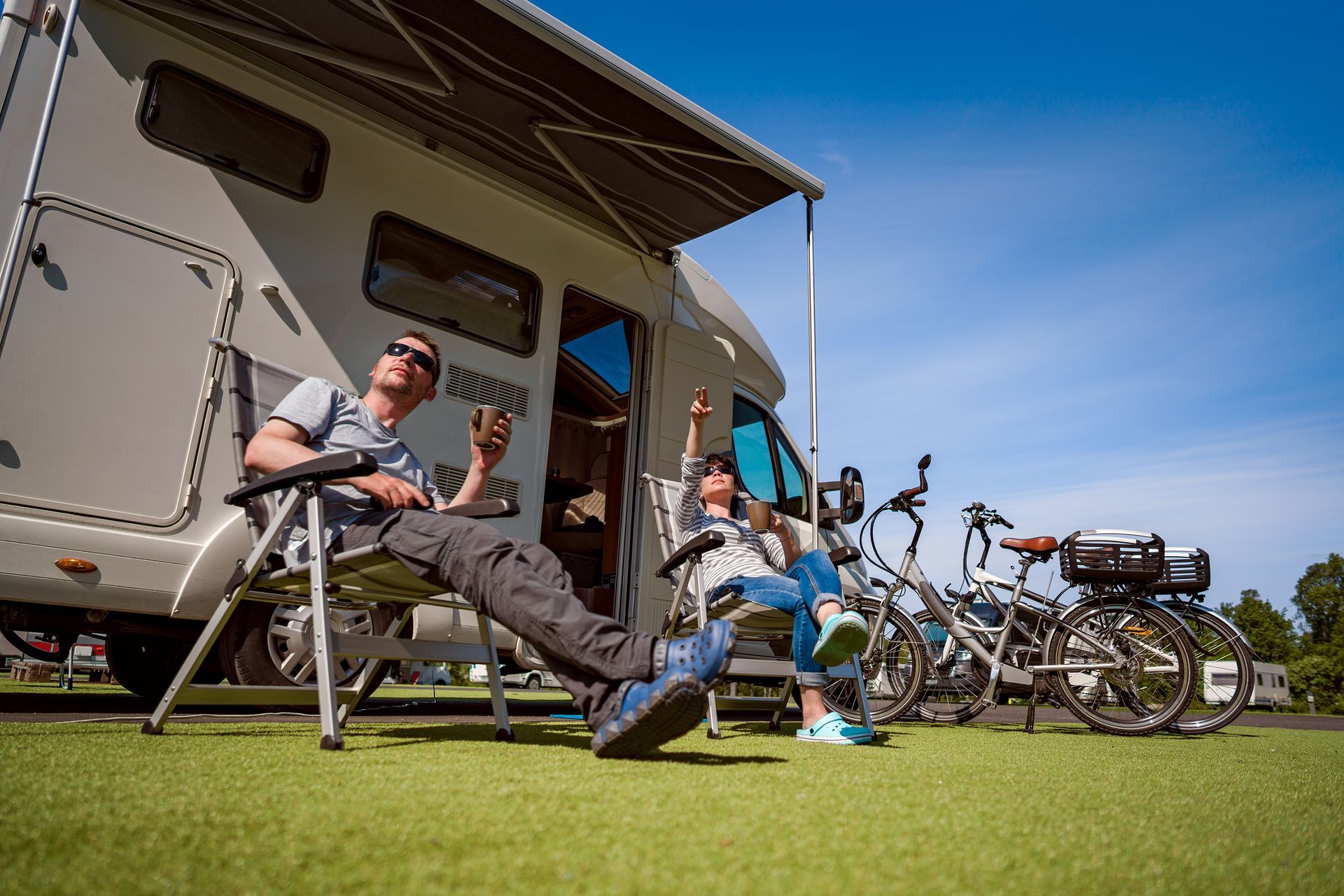 People relaxing in camping chairs beside a camper van and bicycles on a grassy campsite.