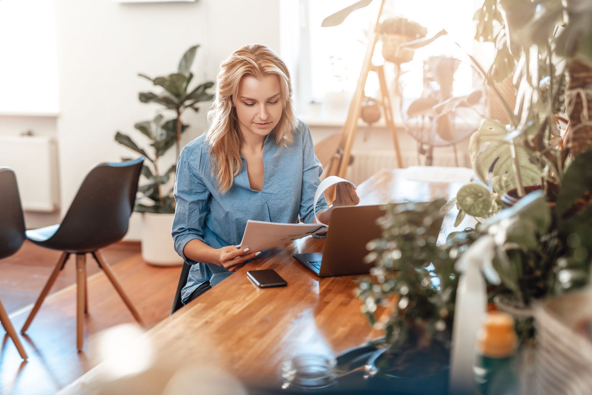 Woman working on a laptop at a wooden desk in a bright home office with plants and a coffee cup.
