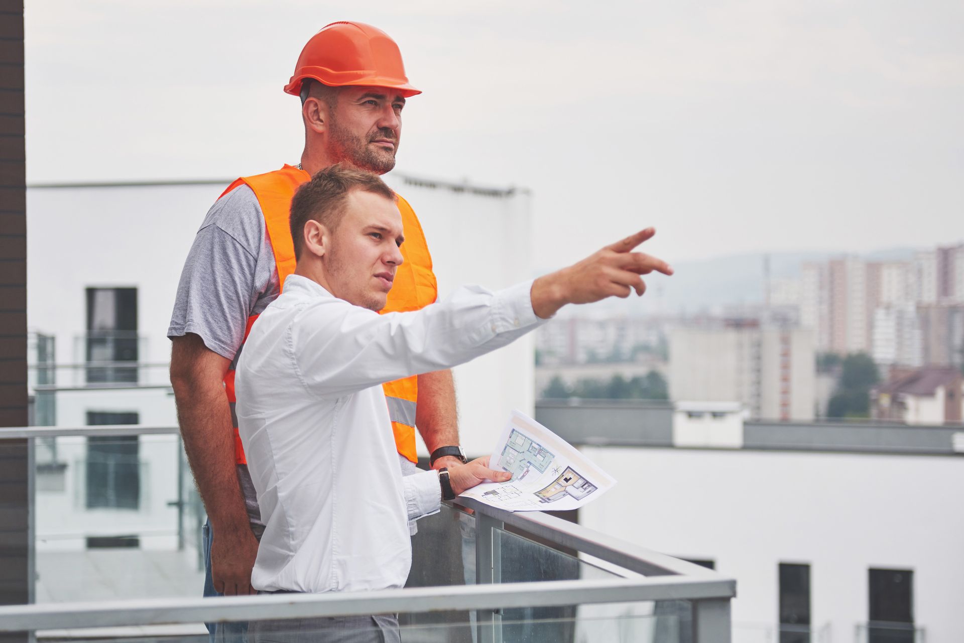 Two workers in safety gear on a rooftop, one pointing over the city skyline