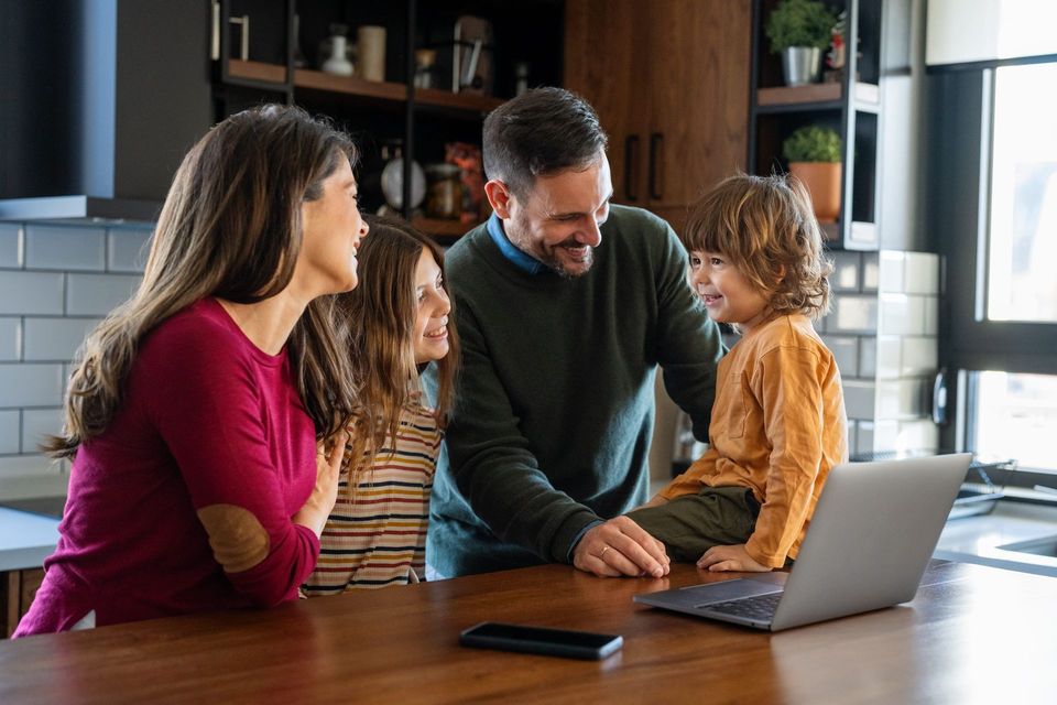 Family gathered around a laptop at a kitchen table, smiling and talking together
