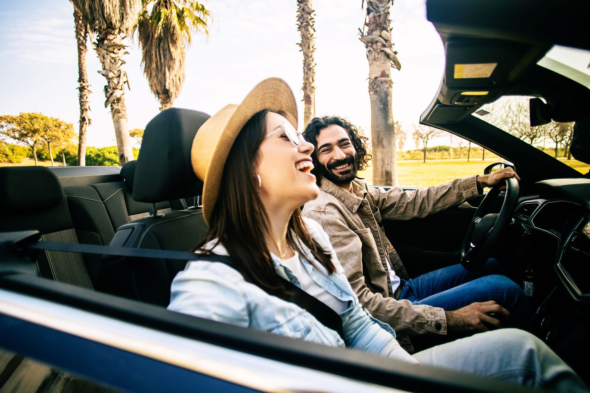 Two people smiling in a convertible with the top down on a sunny road lined with palm trees