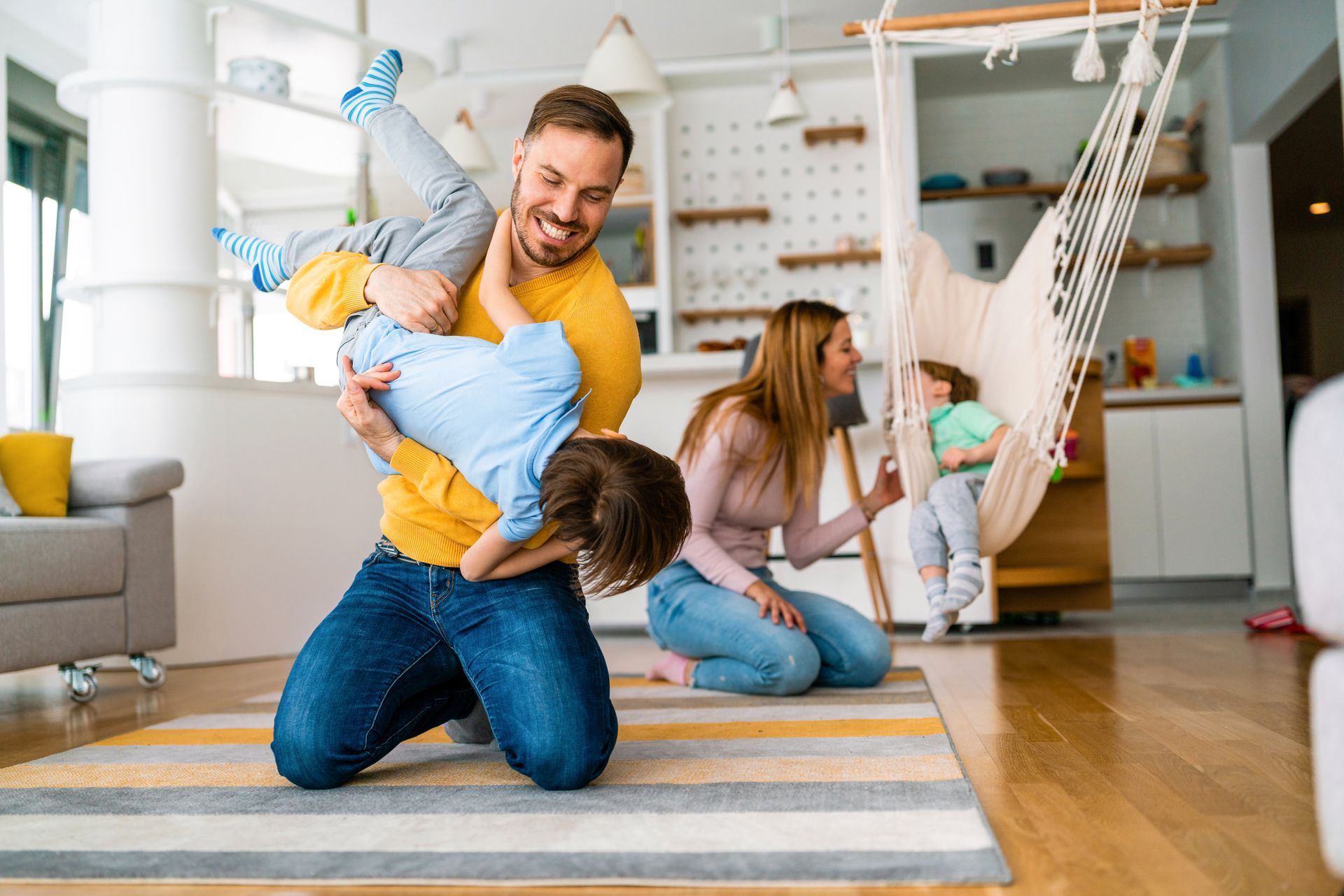 Man kneels with two children playing on a rug in a bright living room with a hammock chair.