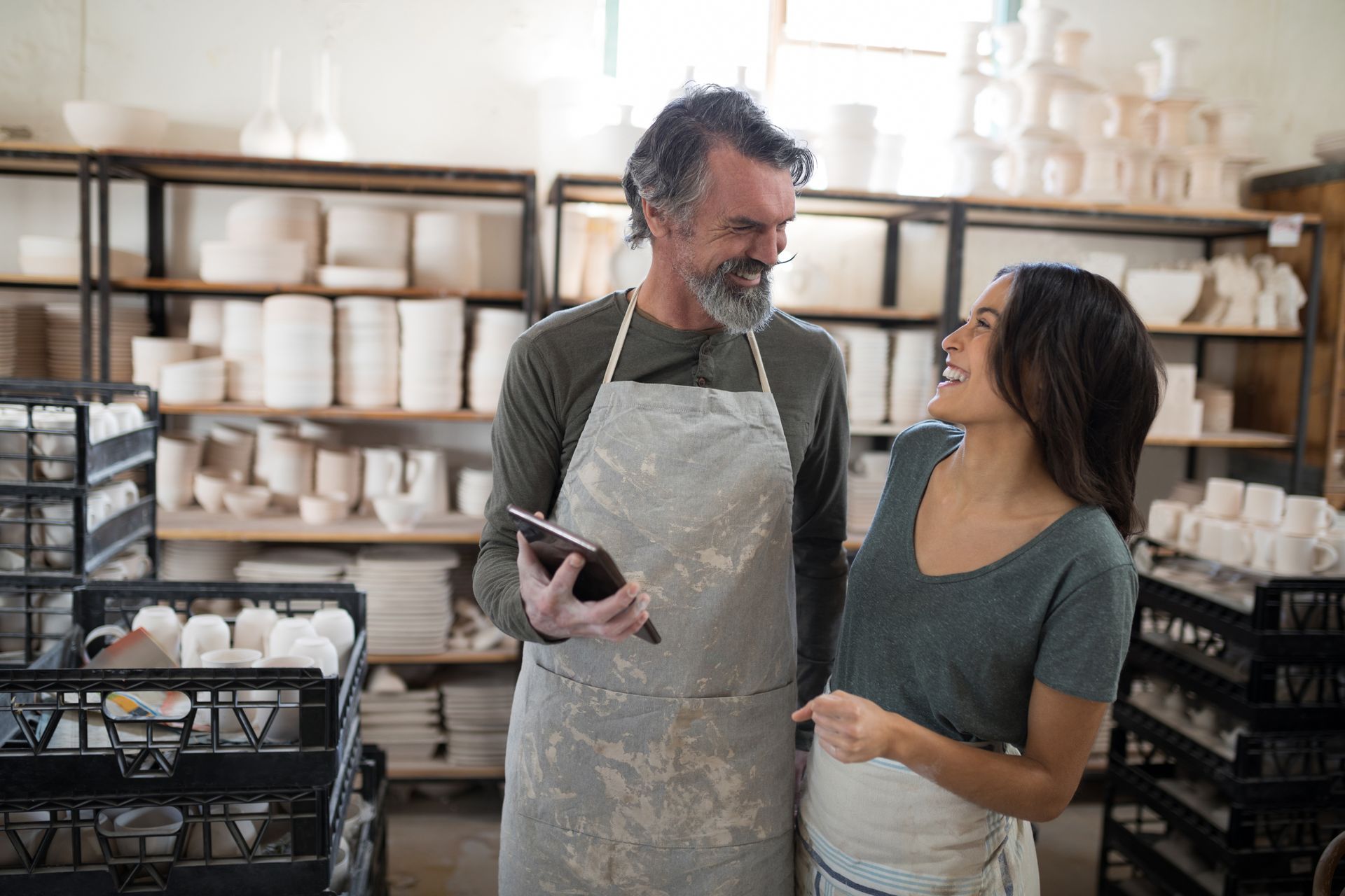 Two people smiling in a ceramics workshop, holding a tablet amid shelves of pottery supplies