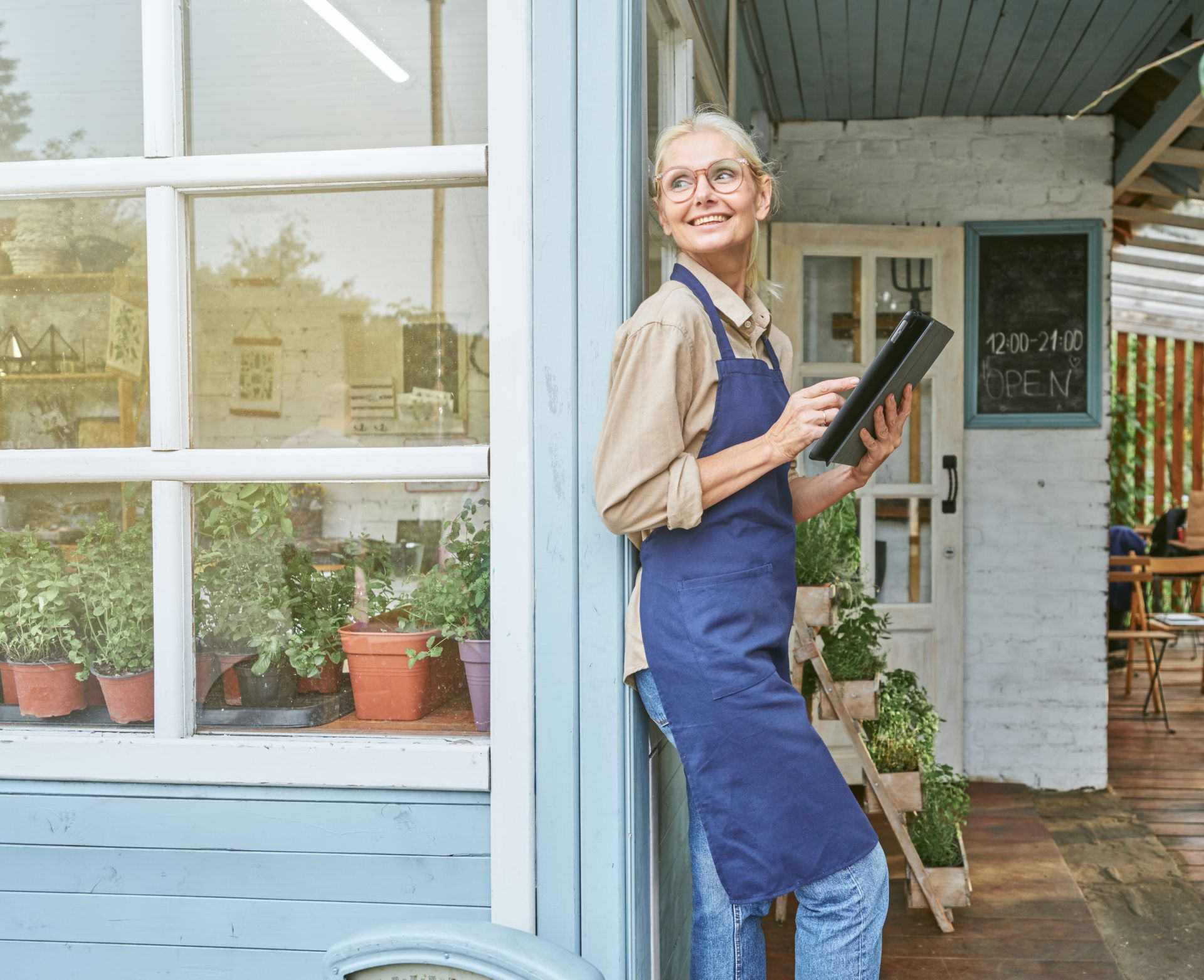 Shopkeeper in blue apron holding a menu outside a storefront, smiling near potted plants.