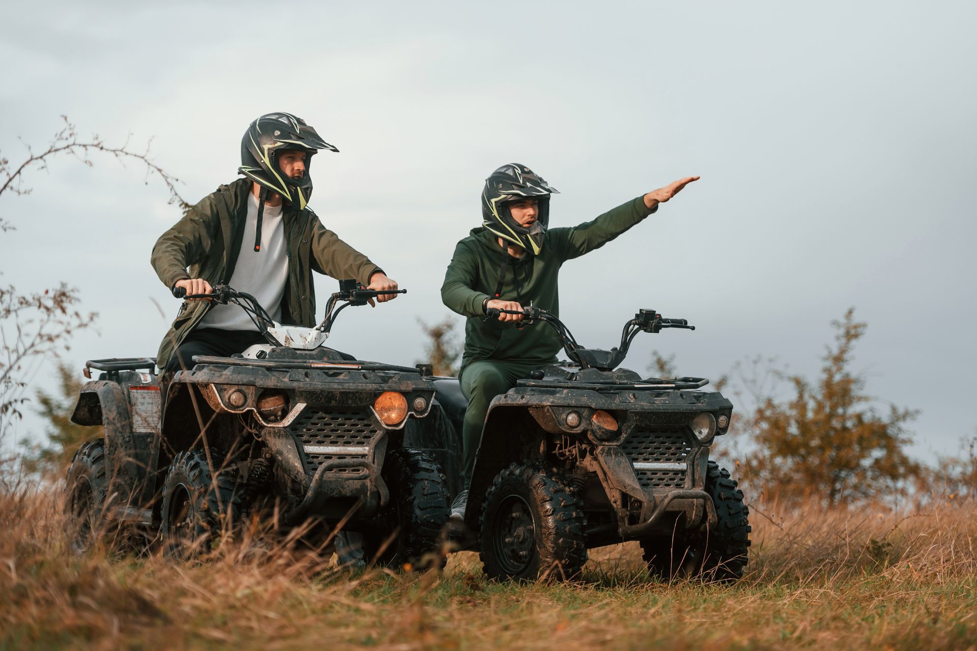 Two helmeted riders on ATVs in a grassy field, one pointing ahead under an overcast sky