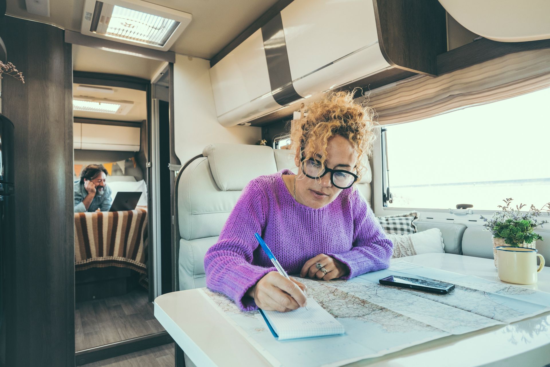 Person in a purple sweater writing at a table inside a modern RV, with sunlight and a phone nearby
