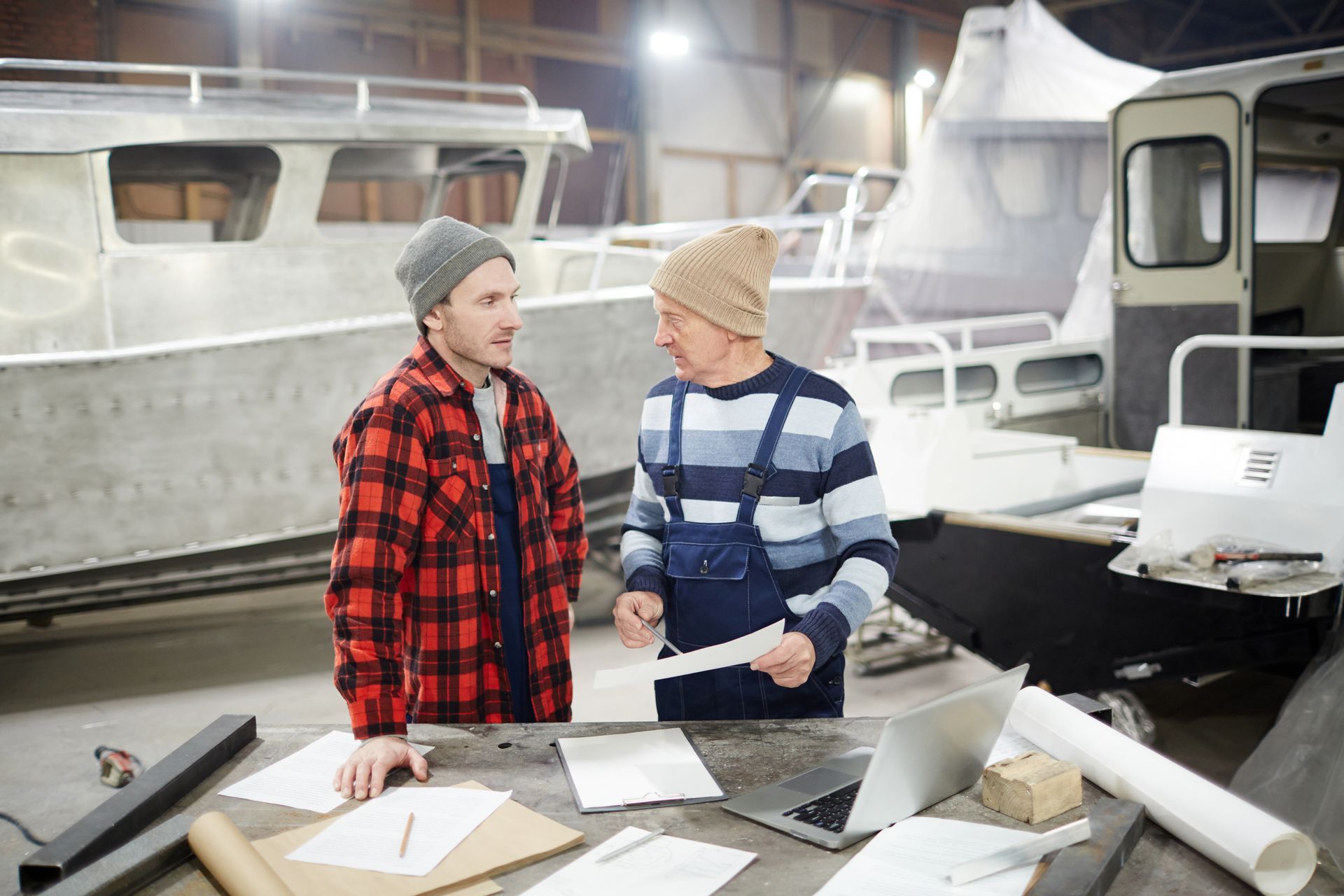 Two workers reviewing blueprints at a table in a boat workshop