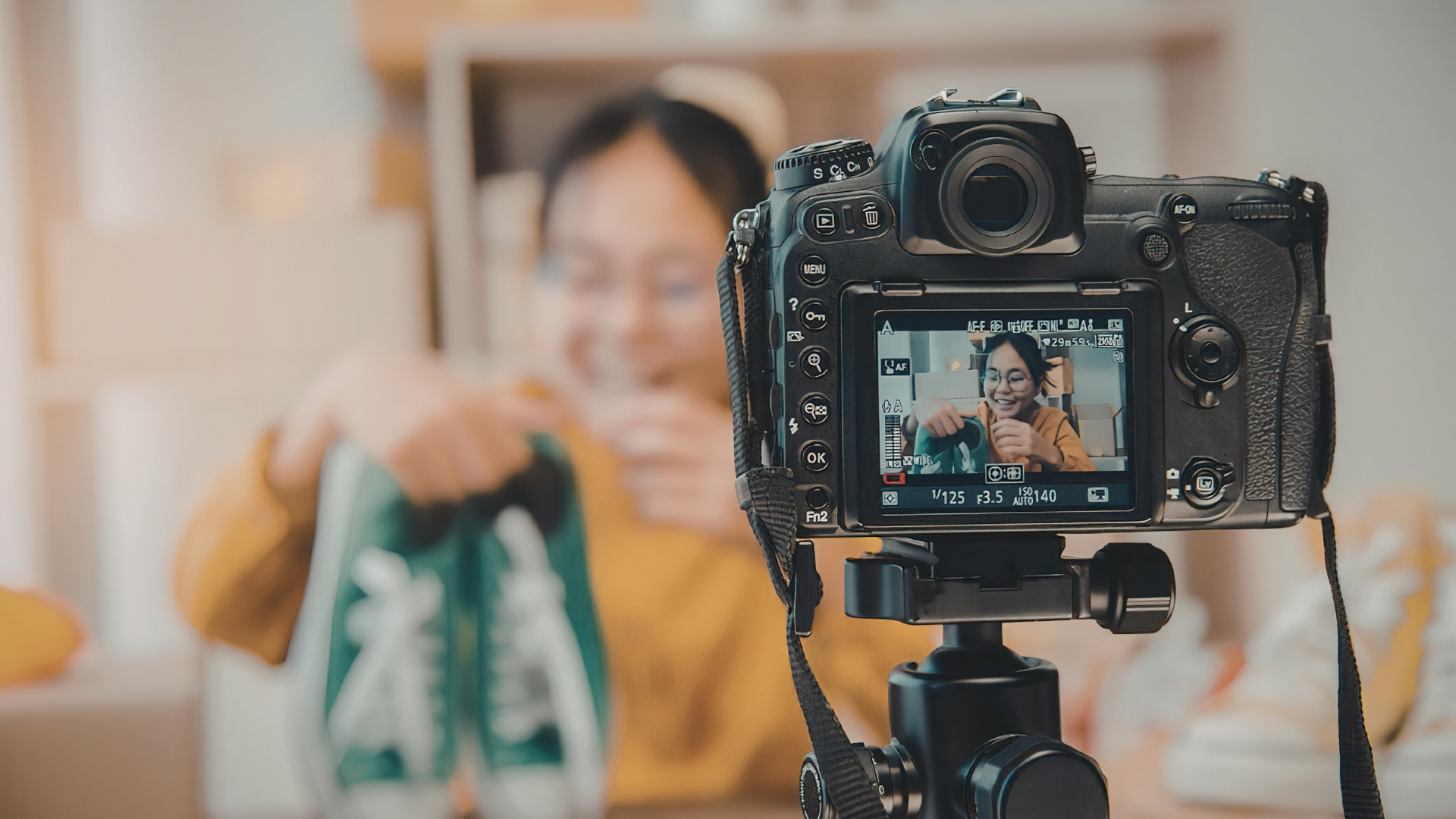 A camera on a tripod faces a person wearing a yellow top who is holding green sneakers for an online video presentation.