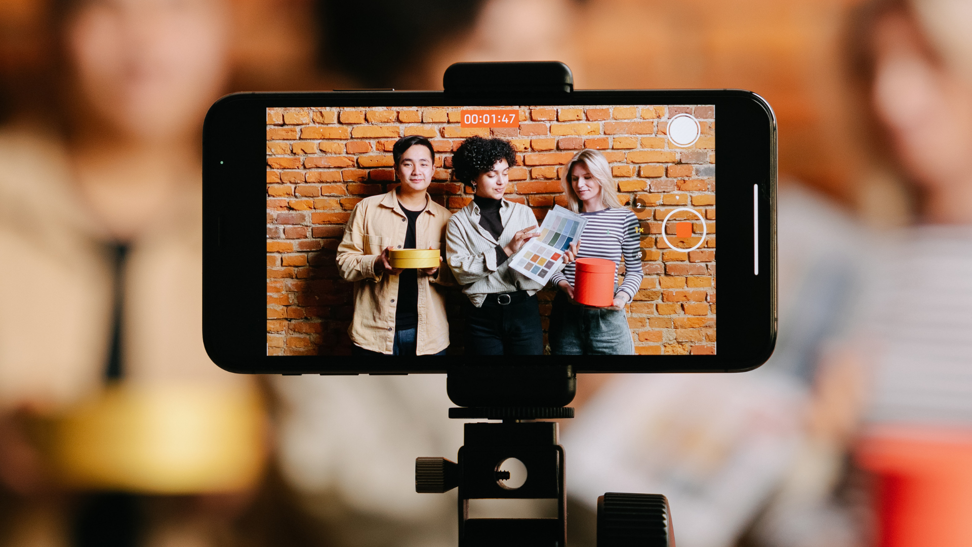A smartphone on a tripod recording three people standing against a brick wall, holding items and showing a document.