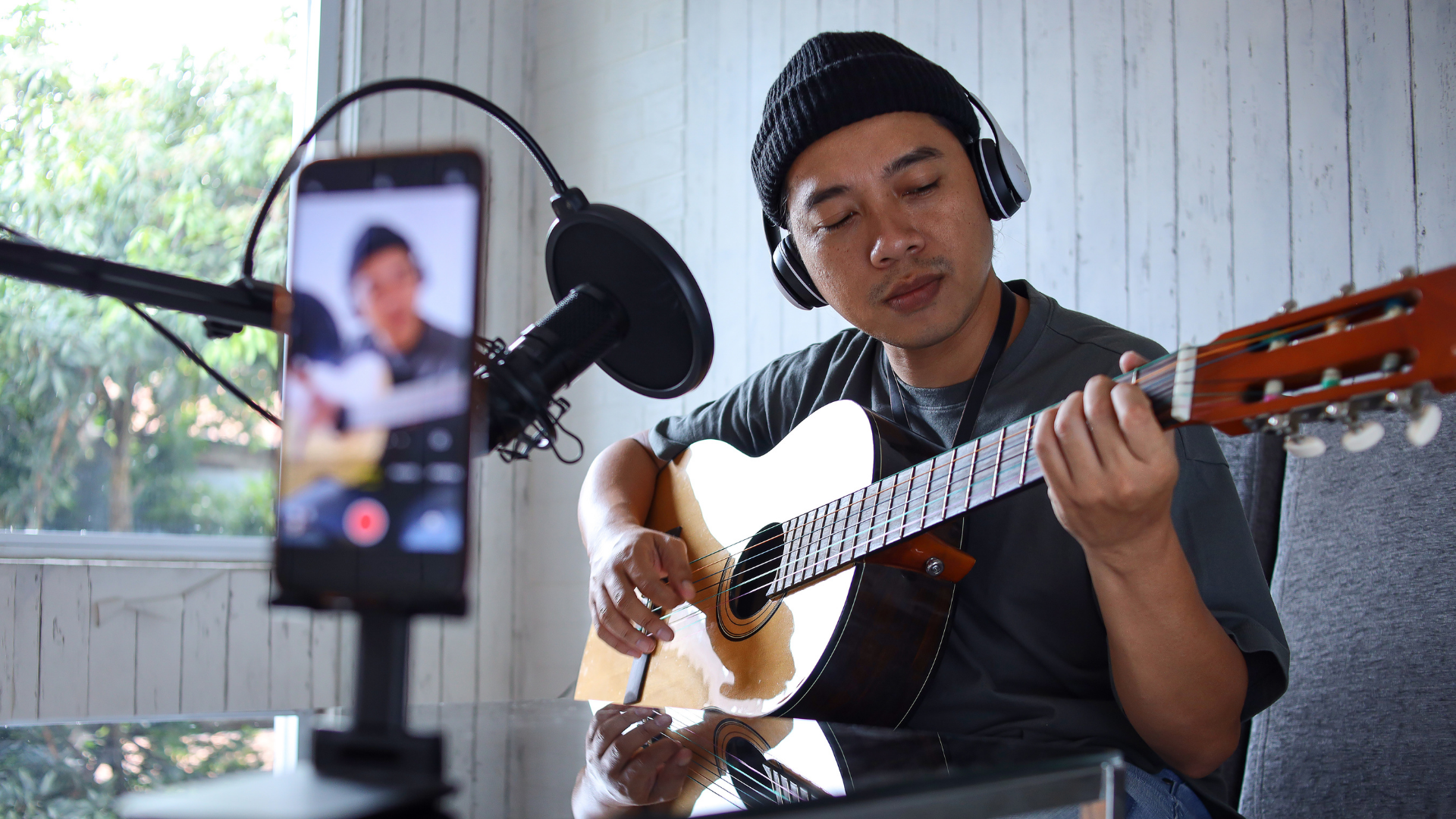 A musician in a beanie and headphones plays an acoustic guitar while recording a video on a smartphone.